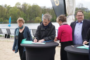 Mehrere Personen stehen um einen Stehtisch an einem Seeufer. Im Zentrum steht Cordelia Koch, sie unterschreibt ein Dokument.