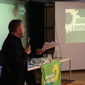 Eine Person spricht vor einem Podium, es ist der EU-Abgeordnete Sergey Lagodinsky. Er hält ein Redemanuskript und ein Mikrofon in der Hand. Im Hintergrund eine Präsentation im Design von B'90/GRÜNE.