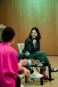 Totale einer jungen Frau mit Schulterlangen Haaren im Gespräch– es ist Julia Schneider. Sie sitzt auf einem Stuhl und hält ein Mikrofon in der Hand. Im Vordergrund der Rücken einer anderen Person mit einer pinken Bluse, im Hintergrund eine Holzvertäfelung, die grün angeleuchtet wird.