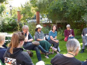 Eine Gruppe von Menschen, die in einem Gesprächskreis in einem Garten sitzen. Im Hintergrund Sträucher und eine alte Gartenmauer.