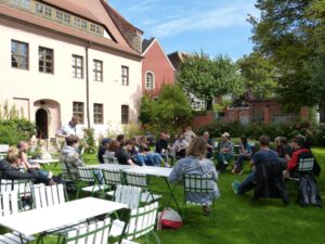 Eine Gruppe von Menschen, Tische und Stühle in einem Garten vor einem Altbau. Die Sonne scheint und Wiese und Hecken sind grün.
