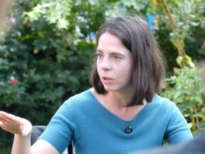 Eine junge Frau mit schulterlangen Haaren im Gespräch – es ist Julia Schneider. Sie trägt ein blaugrünes Oberteil, im Hintergrund Hecken.