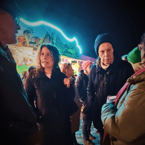 Mehrere Menschen stehen abends auf einem belebten Weihnachtsmarkt im Gespräch. Im Vordergrund diskutieren zwei Personen miteinander, umgeben von weiteren Besucher*innen in Winterkleidung. Im Hintergrund sind ein beleuchteter Fahrgeschäft-Stand und bunte Lichter zu sehen.