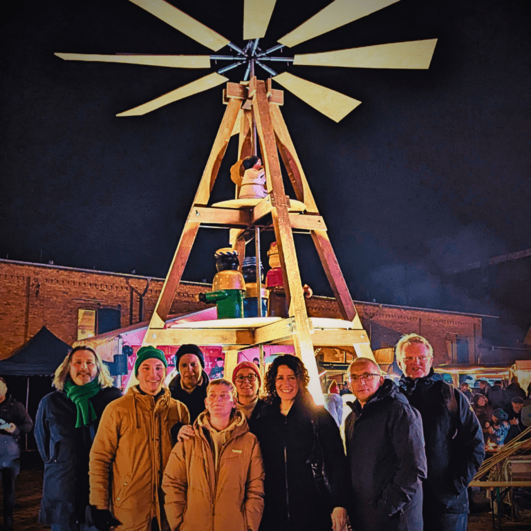Eine Gruppe von Menschen steht abends auf einem Weihnachtsmarkt vor einer großen, beleuchteten Holzpyramide. Alle tragen warme Winterkleidung. Im Hintergrund sind Marktstände, Lichter und weitere Besucher*innen zu sehen.