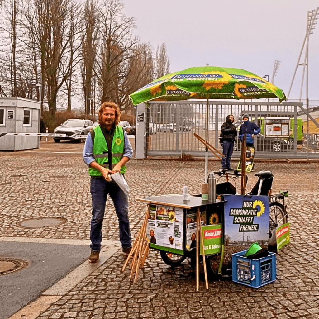 Ein Mann mit grüner Warnweste steht neben einem Lastenrad, das als Infostand genutzt wird. An dem Rad hängen Plakate, Greifzangen und ein Sonnenschirm. Im Hintergrund ist ein eingezäunter Platz mit weiteren Personen.