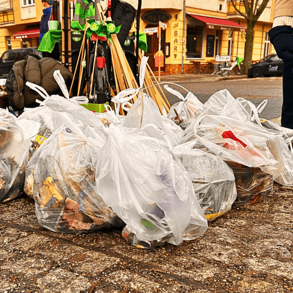 Mehrere durchsichtige Müllsäcke voller Abfall stehen auf Kopfsteinpflaster. Dahinter sind Greifzangen und grüne Windräder an ein Lastenrad angelehnt. Menschen und Häuser sind unscharf im Hintergrund.