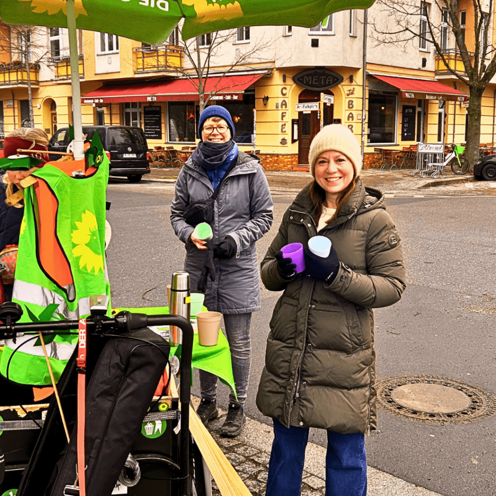Zwei Frauen stehen an einem Lastenrad mit grünem Sonnenschirm. Sie tragen Winterjacken und Mützen und halten Becher in den Händen. Im Hintergrund sind Häuser, ein Café und parkende Autos zu sehen.