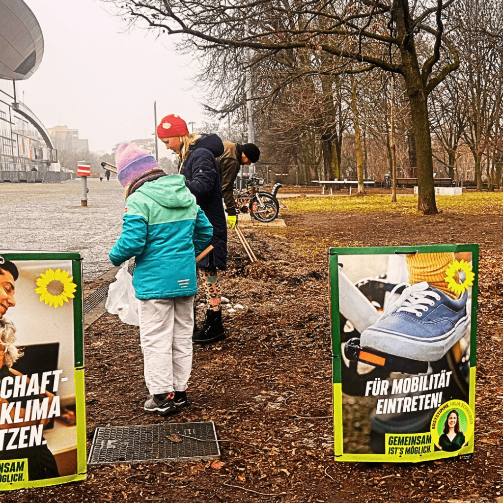 Mehrere Personen, darunter ein Kind, sammeln Müll auf einem Weg im Park. Sie tragen Winterjacken und Mützen und benutzen Greifzangen. Im Vordergrund stehen zwei grüne Wahlplakate am Wegesrand.