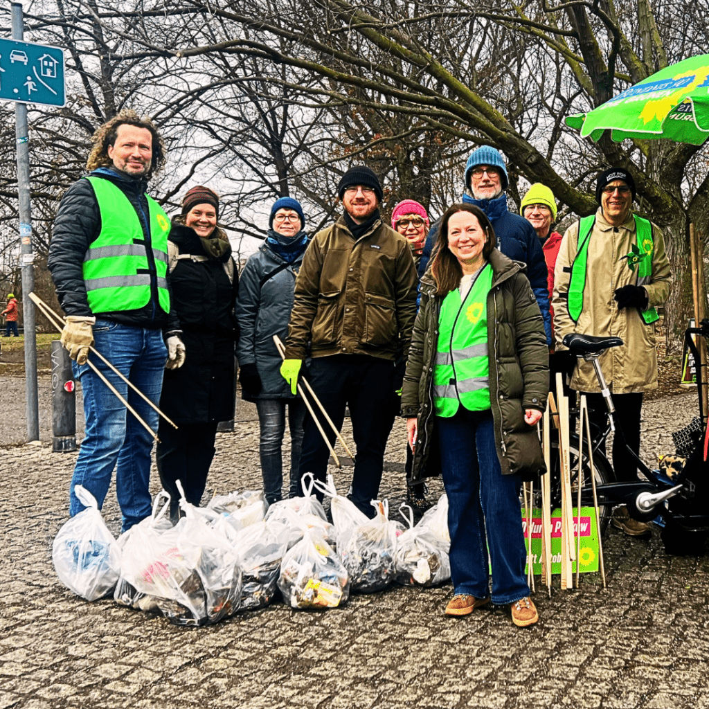 Eine Gruppe von etwa zehn Menschen steht im Freien auf einem Platz mit Bäumen. Sie tragen Winterkleidung und grüne Warnwesten. Vor ihnen liegen mehrere gefüllte Müllsäcke. Einige halten Greifzangen. Alle schauen in die Kamera.