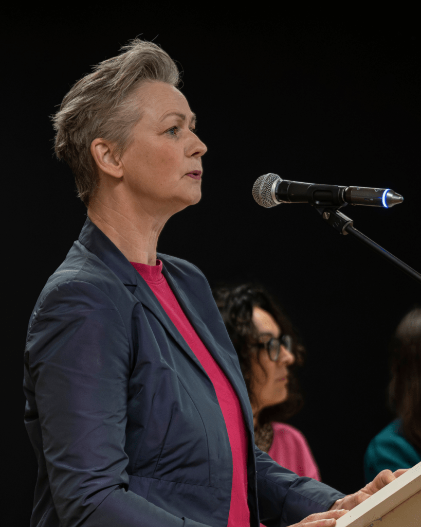 Eine Frau mit kurzen Haaren steht an einem Mikrofon und spricht. Es ist die Pankower Bürgermeisterin Cordelia Koch. Sie trägt ein dunkles Sakko und ein pinkes Oberteil. Im Hintergrund sitzen weitere Personen auf der Bühne.