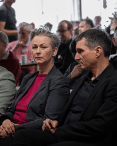 Eine Frau mit kurzen Haaren und ein Mann sitzen nebeneinander im Publikum und schauen nach vorne. Es sind Cordelia Koch und Cornelius Bechtler. Hinter ihnen sind weitere Zuhörende in einem Veranstaltungsraum zu sehen.