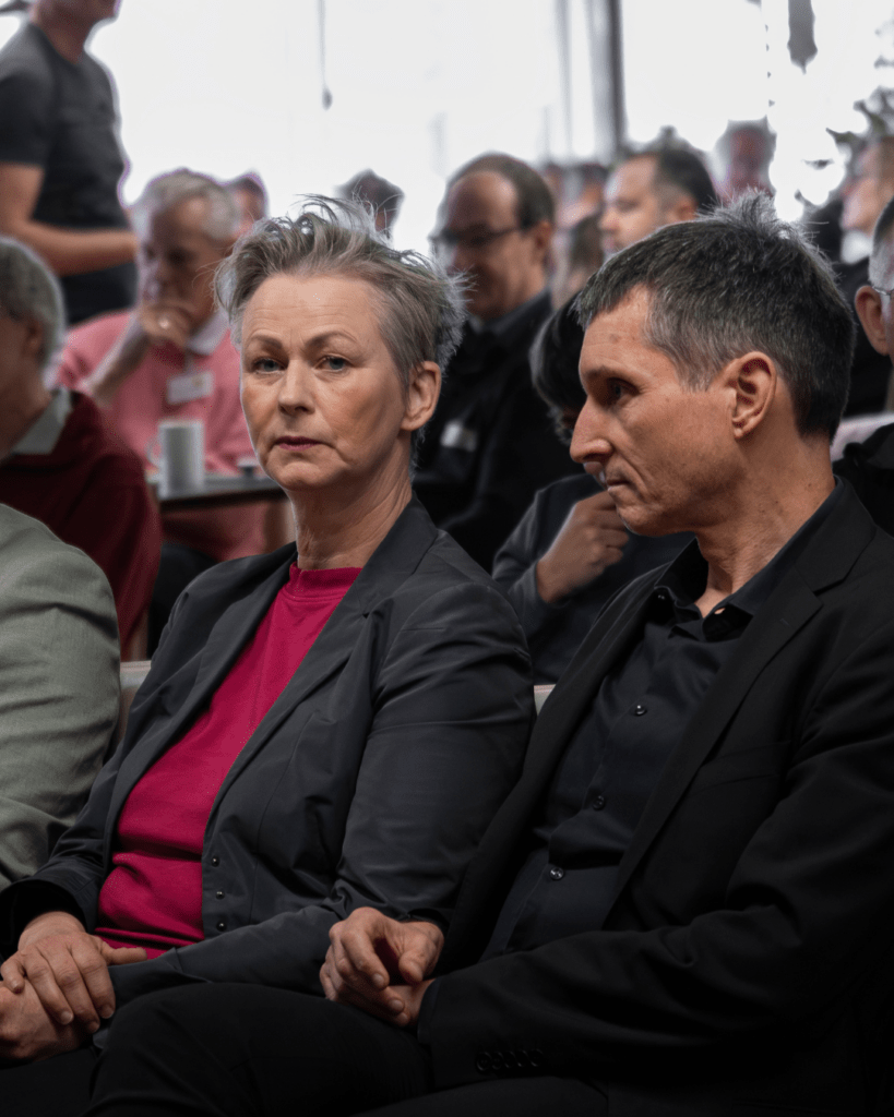 Eine Frau mit kurzen Haaren und ein Mann sitzen nebeneinander im Publikum und schauen nach vorne. Es sind Cordelia Koch und Cornelius Bechtler. Hinter ihnen sind weitere Zuhörende in einem Veranstaltungsraum zu sehen.