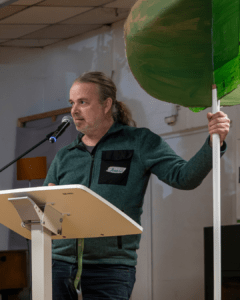 Ein Mann steht an einem Redepult mit Mikrofon und hält ein großes, grün bemaltes Schild in Form eines Baumes. Er spricht vor Publikum in einem Veranstaltungsraum.