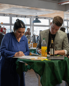 Eine Frau und ein junger Mann stehen an einem runden Tisch mit grüner Tischdecke und schreiben. Es sind Paul Schlüter und Almuth Tharan. Auf dem Tisch stehen Getränke, ein Laptop und ein Teller mit Keksen. Im Hintergrund sind weitere Personen im Raum zu sehen.