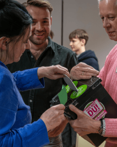 Mehrere Personen stehen dicht beieinander und werfen Stimmzettel in eine schwarze Wahlurne. Auf der Urne klebt ein Aufkleber mit der Aufschrift „Hier bist DU entscheidend“.